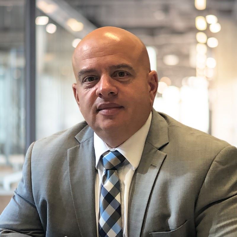 Headshot of a man in a grey suit with a blue plaid tie
