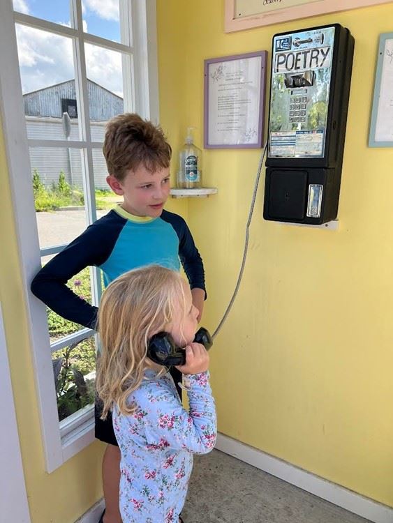 Young girl in a floral shirt listens to a payphone with older boy looks on